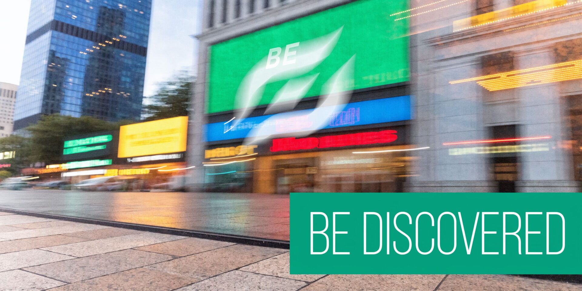 A motion-blurred city street scene in front of a modern building featuring a prominent be discovered sign.