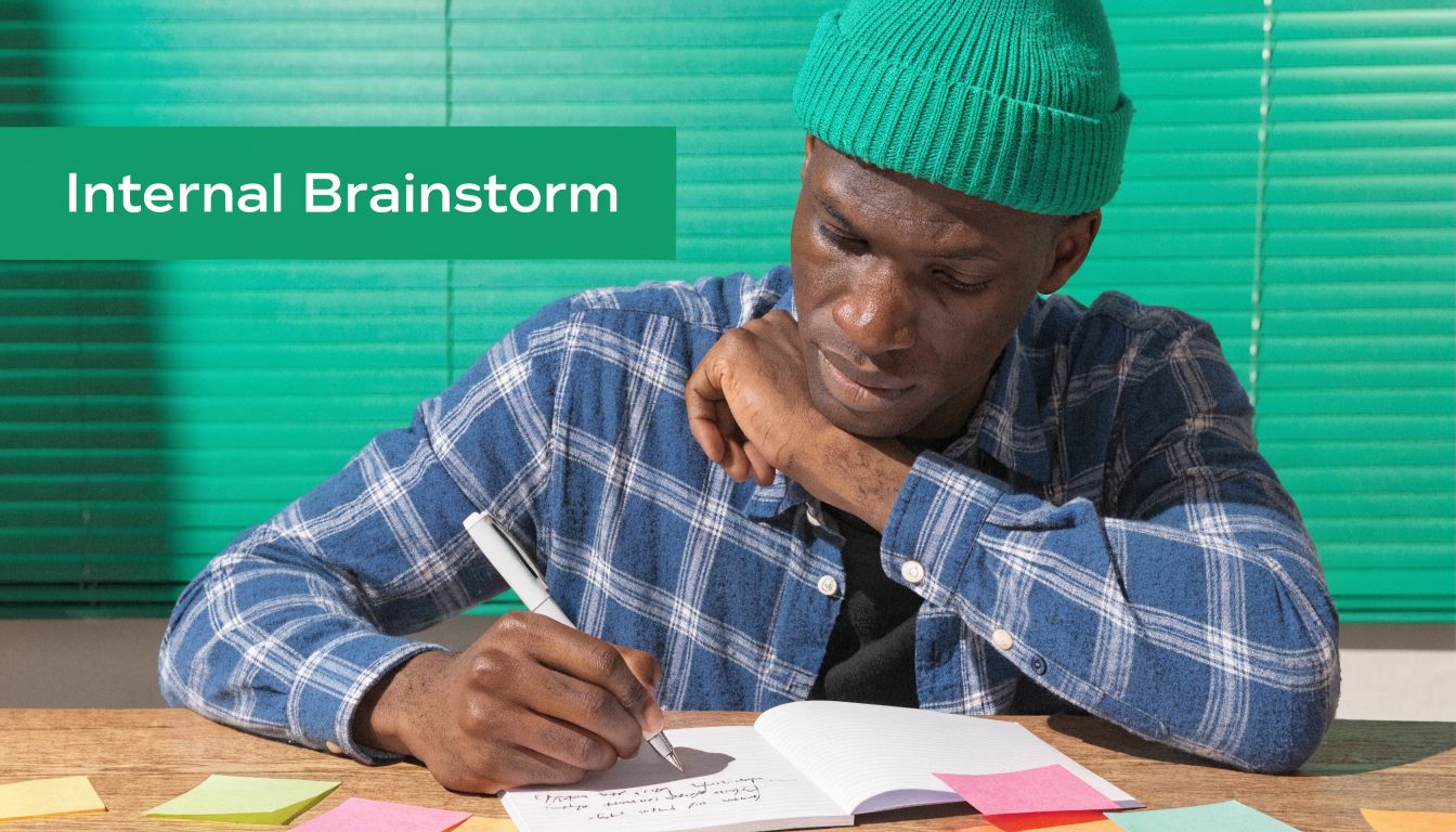 A focused man wearing a green beanie writing in a notebook during an internal brainstorming session.